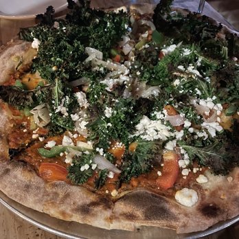 Reno - Alley-Oop - A pizza with kale, vegetables, and cheese is served on a metal tray on a wooden table, with a white plate and glass in the background. - American Near Me - Chicago, Illinois
