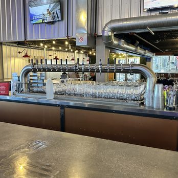 Rio Bravo Brewing - tap wall - A bar counter with a row of beer taps, glasses, and bar tools, inside a brewery or pub with industrial decor and large windows. - American Near Me - Albuquerque, New Mexico