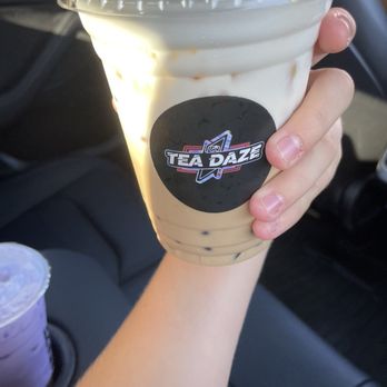 Tea Daze - Sea salt coffee with lychee jelly - A hand is holding a large plastic cup of tea latte with tapioca pearls, branded with the Tea Daze logo. The drink has a creamy appearance with visible tapioca pearls at the bottom. The background shows the interior of a vehicle, indicating the drink was purchased for on-the-go consumption. - Coffee & Tea Near Me - Modesto, California