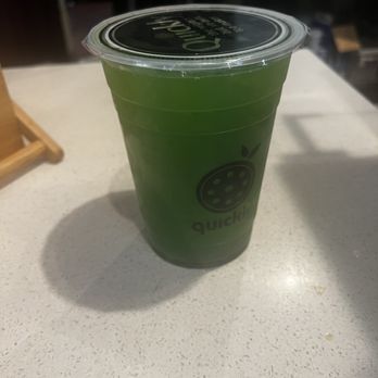 Quickly - Ann Arbor - Kiwi lavender fruit tea - A green fruit tea drink in a clear plastic cup with a black lid, featuring a logo with a fruit graphic and the word 'quick'. The drink is on a light-colored countertop, with a shadow cast to the left. The background shows a blurred cafe environment with equipment and shelves. - Bubble Tea Near Me - Ann Arbor, Michigan