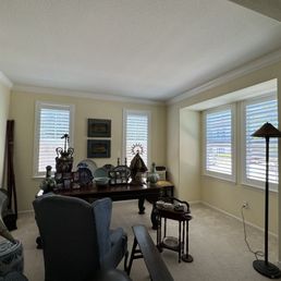 New shutters - The image shows a living room with three windows fitted with white blinds, allowing natural light to enter. There is a wooden dining table with decorative items, including vases and framed photos, positioned against the wall. The room contains upholstered chairs, a floor lamp, and other furniture, with a neutral-colored carpet and light-colored walls.