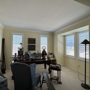 SEI Window Coverings - New shutters - The image shows a living room with three windows fitted with white blinds, allowing natural light to enter. There is a wooden dining table with decorative items, including vases and framed photos, positioned against the wall. The room contains upholstered chairs, a floor lamp, and other furniture, with a neutral-colored carpet and light-colored walls. - Shutters Near Me