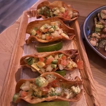 Bien Trucha - Four crispy taco shells filled with diced vegetables including avocado, tomato, and onion, are arranged on a wooden serving board with lime wedges placed underneath. The tacos are garnished with herbs and appear to contain additional toppings like cheese or sauce. The setting suggests a restaurant environment with a wooden table and a bowl of similar food nearby. - Mexican Near Me - Wheaton, Illinois