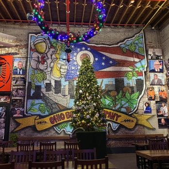 Ohio Brewing - Sunday funday at OBC! - A Christmas tree decorated with lights and ornaments is positioned in the center of a restaurant interior. Behind it, a mural on the brick wall depicts a cartoon astronaut holding a beer bottle, with a flag featuring a cityscape and hop plants. A large, illuminated holiday wreath hangs from the ceiling above the tree. The restaurant has wooden tables and chairs, and framed photographs are displayed on the walls. - Breweries Near Me - Columbus, Ohio