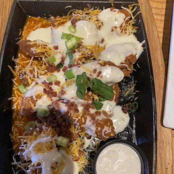 Chili's - Fried chicken tenders are arranged on a black serving tray, topped with shredded cheese, chopped green onions, and drizzled with a creamy white sauce. A small cup of additional creamy sauce is placed on the tray. The tray is set on a wooden table, and the image captures the dish from a top-down perspective. - American Near Me - Williston, Vermont