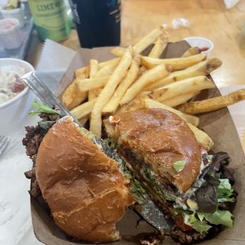 Sacred Ground - Smash burger and fries - A burger with a toasted bun, lettuce, tomato, and sauce is cut in half and placed on a tray with a portion of French fries. The fries are golden and slightly crispy. The background shows drinks and condiments on a wooden table, indicating a casual restaurant setting. - Salad Near Me - Flora, Mississippi