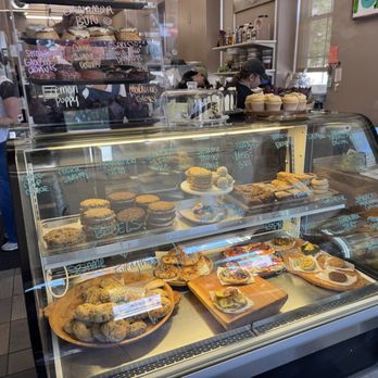 Harmony Bakery - Display case of baked goods - A glass display case showcases various baked goods including cookies, scones, and pastries, with handwritten labels on the glass indicating the items. Behind the case, bakery staff and customers are visible, some preparing or serving food. The bakery interior features shelves with supplies and a small seating area near windows, creating a cozy atmosphere. - Gluten-Free Near Me - Baltimore, Maryland