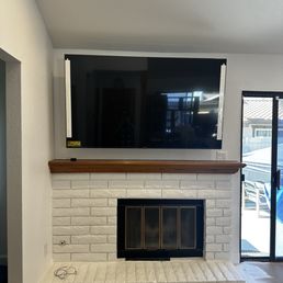 A flat-screen TV is mounted above a white brick fireplace with a wooden mantel in a living room, with a sliding glass door to the right.