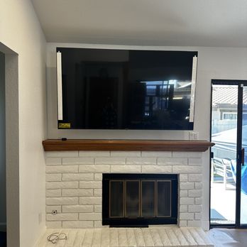 99 TV Install - A flat-screen TV is mounted above a white brick fireplace with a wooden mantel in a living room, with a sliding glass door to the right. - Home Theatre Installation Near Me - Upland, California