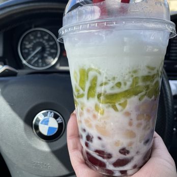 BAMBU - Che 3 mau (wouldn't get again) - A bubble tea drink with green jelly, tapioca pearls, and milk is held in front of a BMW steering wheel inside a vehicle. - Bubble Tea Near Me - Minneapolis, Minnesota
