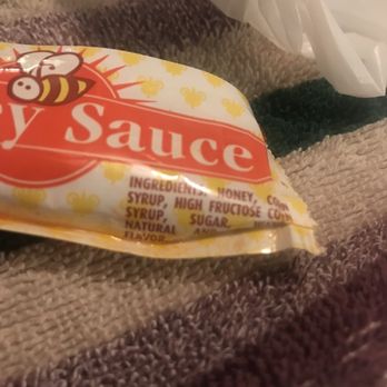 Casa De Benavidez - A small, colorful candy packet labeled 'Honey Sauce' features a cartoon bee illustration and a background with yellow honeycomb patterns. The ingredients listed include honey, corn syrup, high fructose corn syrup, sugar, and natural flavor. The packet is resting on a textured fabric surface, with a white plastic bag partially visible in the background. - Mexican Near Me - Albuquerque, New Mexico