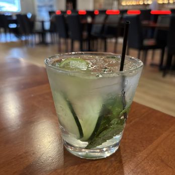 Vortex Eatery Plus Taphouse - Gin &Tonic - A clear glass contains a gin and tonic with lime slices and fresh mint leaves, filled with ice and a straw. The drink is placed on a wooden table in a restaurant or bar environment, with blurred background seating and decor visible, including chairs and shelves with bottles or glasses. - Burgers Near Me - Salem, Oregon