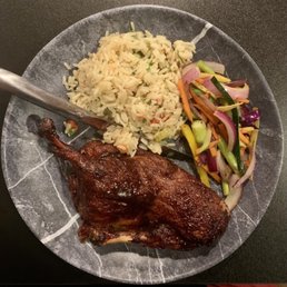 Duck Confit - A plate contains rice, a grilled chicken leg, and a vegetable salad with sliced cucumbers, carrots, and red onions.