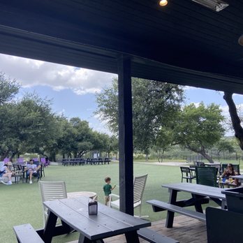 Frontyard Brewing - Sitting area - Outdoor seating area at Frontyard Brewing features picnic tables and chairs under a covered patio, with a view of a grassy field, trees, and a partly cloudy sky. People are sitting at tables, some with children, and a few are standing or walking. The area includes a playground or recreational space in the background, with a few individuals engaged in activities. The scene depicts a family-friendly, community-oriented outdoor space. - Breweries Near Me - Spicewood, Texas