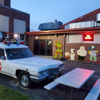 Old School Pizza Malta - Front door - A brick pizza restaurant features cartoon characters from 'Family Guy' on the wall and a neon sign. A Ghostbusters-themed car is parked outside near a picnic table. - Pizza Near Me - Malta, Illinois