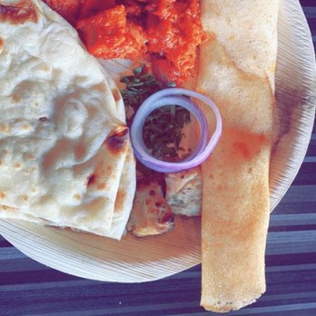 India 101 - Chicken 65 - A round plate contains Indian dishes including naan bread, spicy chicken 65 with red sauce, a crispy papadum, and a small container of green chutney. The plate is placed on a dark striped table, with the restaurant name and location, India 101 in Irving, TX, visible at the bottom of the image. - Indian Near Me - Irving, Texas