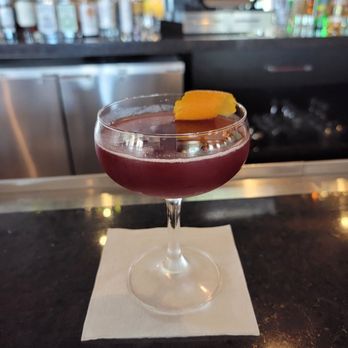 Zorba Lounge - Fig-Syrup'R - A dark red cocktail in a coupe glass with an orange peel garnish, placed on a bar counter with a bar and cash register in the background. - Lounges Near Me - Naperville, Illinois