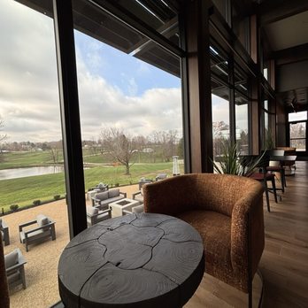Rhonemus Cellars - Dining room - The image shows the interior of a modern winery with expansive glass windows providing a view of a golf course, trees, and a pond outside. Inside, there are wooden floors, a brown upholstered chair, a small black wooden table, and some potted plants. Additional seating with chairs and tables is visible further inside, emphasizing a spacious and contemporary setting suitable for wine tastings and relaxing small bites. - Wineries Near Me - Lancaster, Ohio