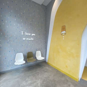Machi Machi - The image shows a seating area with three chairs, two white and one olive green, positioned against a wall decorated with colorful sprinkles and a message that reads, "I love you so machi." The floor appears to be concrete, and the space has a modern, minimalistic design with a curved yellow wall on the right side. The lighting is soft, with a small spotlight illuminating the wall with the message. - Bubble Tea Near Me - Markham, Ontario