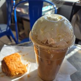 Iced Caramel Machiatto - A caramel frappuccino with whipped cream on top is in a clear plastic cup with a domed lid, sitting on a table. Next to it, there is a piece of fried chicken on a white napkin. The background shows a window with sunlight and some chairs, indicating a casual dining or coffee shop setting.