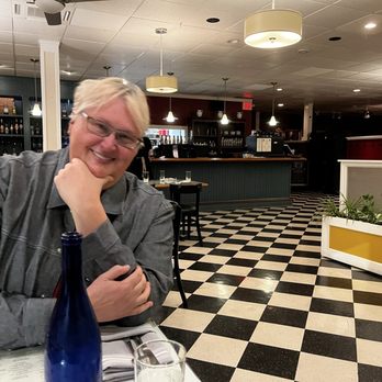 Le Central - A person with glasses and blonde hair, wearing a gray shirt, smiling at a restaurant table with a blue bottle and glass in front. - French Near Me - Bristol, Rhode Island