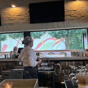 NiMo - A person in a white shirt, likely a bartender, stands behind a bar counter with various glassware, bottles, and bar tools. The background features a large window with a view of trees and a mural or painted wall outside. The interior has a brick wall with mounted screens and warm lighting. - Salad Near Me - Tequesta, Florida