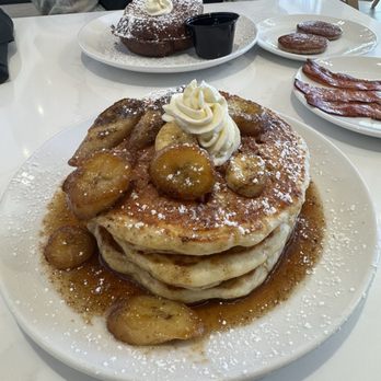 Brunch & Sip - Banana foster pancakes - A plate of thick pancakes topped with caramelized bananas and whipped cream, served with syrup at a brunch spot with additional breakfast items in the background. - Coffee & Tea Near Me - Phoenix, Arizona
