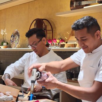 Two Nine - Two chefs in white uniforms prepare sushi at a counter, with decorative plates, flowers, and a ukulele on shelves behind them. - Sushi Bars Near Me - Washington, DC, District of Columbia