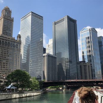 Chicago Architecture Center - Initial voyage toward the river branch. - A cityscape of Chicago with numerous tall skyscrapers lining the riverbank. The buildings vary in architectural style, with some featuring glass facades and others with traditional designs. A bridge crosses the river in the foreground, and a few people are visible at the bottom of the image, indicating a boat cruise or river activity. The sky is partly cloudy, and the scene captures the urban environment and architectural diversity of Chicago. - Historical Tours Near Me - Chicago, Illinois