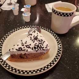 Chocolate cream pie and coffee - A slice of chocolate cream pie topped with whipped cream and chocolate sprinkles is placed on a checkered-pattern plate. A metal pie server rests under the pie. Next to the plate, there is a white ceramic mug filled with coffee. The table surface is dark and speckled, with various small containers and utensils visible in the background, indicating a diner setting.