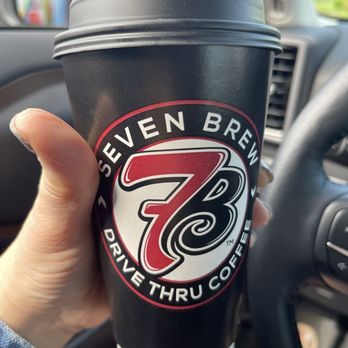 7 Brew Coffee - A black disposable coffee cup from Seven Brew Coffee with a lid, held by a person inside a vehicle, with the vehicle's dashboard and steering wheel visible. - Coffee & Tea Near Me - Springfield, Missouri