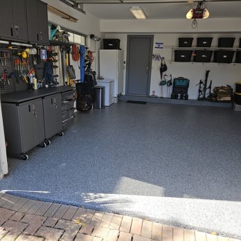 Garage Store - new garage floor - A well-maintained garage features dark gray cabinets and a pegboard with various tools hanging on the left wall. The garage has a white refrigerator, a small white cabinet, and a white refrigerator near the back wall. Storage bins are placed on a wall-mounted shelf, and outdoor equipment such as a lawn mower and garden tools are stored on the right side. The floor is coated with a speckled gray epoxy finish, and the garage door is open, revealing a brick driveway outside. - Flooring Near Me - East Dundee, Illinois