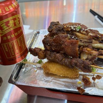 Mala Siblings - Chicken Breast - The image shows a metal tray lined with aluminum foil, containing skewers of grilled meat topped with sesame seeds. There is also a fried item on the tray. A red canned beverage is placed next to the tray. The setting appears to be a casual dining environment, possibly a hot pot or skewer restaurant, with a reflective surface and chopsticks visible in the background. - Specialty Food Near Me - Flushing, New York