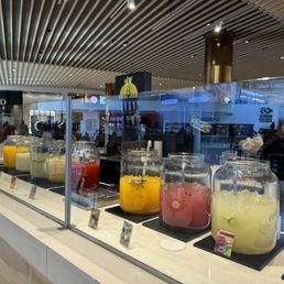 A juice bar in a mall food court offers a variety of fruit-based drinks in large glass jars, with a digital menu and customers in the background.