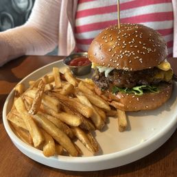 A cheeseburger with a sesame seed bun, lettuce, pickles, and melted cheese, accompanied by a portion of French fries and a small container of ketchup on a white plate. The meal is set on a wooden table, with a person wearing a pink and white striped shirt and a light cardigan visible in the background.