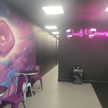 Dollar Donut Shops - Interior of a donut shop with a neon sign 'Donuts & Dreams' on a dark wall, purple chairs, a mural of a pink donut on a galaxy-themed background, and a small seating area. - Donuts Near Me - Howell, New Jersey