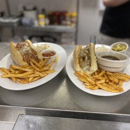 Italian beef and meatball sub - Two white plates contain hot dogs topped with meat and mushrooms, accompanied by French fries. Each plate has small bowls of condiments, including ketchup, mustard, and possibly relish or onions. The background shows a kitchen environment with shelves and containers, and a person wearing a black shirt is partially visible.