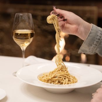 Grace Restaurant - Hand lifting a forkful of long pasta strands above a white bowl, with a glass of white wine and warm candlelight in the background - Diners Near Me - State College, Pennsylvania Grace Restaurant - Hand lifting a forkful of long pasta strands above a white bowl, with a glass of white wine and warm candlelight in the background - Diners Near Me - State College, Pennsylvania