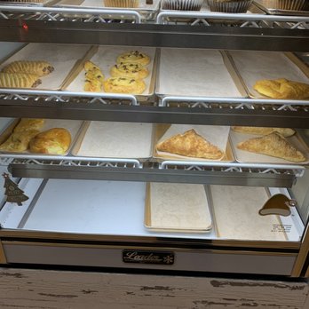 Brooklyn Bread Cafe - The image shows a refrigerated display case with multiple shelves holding baked goods such as muffins, cookies, and pastries. Some trays are empty, indicating items may have been sold or not yet stocked. A small Christmas sign is visible on the top shelf, suggesting a holiday theme. The case is situated on a wooden floor, and the bakery items are arranged on parchment-lined trays. - Sandwiches Near Me - Brooklyn, New York