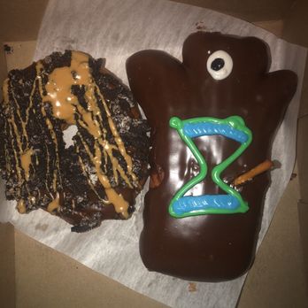 Voodoo Doughnut - Old Dirty Bastard & Voodoo Doll - A bear-shaped cake covered in chocolate icing, featuring a white and black googly eye and a green and blue '2' decoration made of icing or plastic. Next to the cake is a cookie topped with caramel drizzle and crushed cookie pieces. The items are placed on a piece of parchment paper inside a cardboard box, with a plain background. - Donuts Near Me - Denver, Colorado