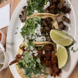 Pastor Taco - A white oval plate holds three tacos filled with different types of grilled meats, topped with chopped onions and cilantro. Two lime wedges are placed on the side of the plate. The tacos are served on a wooden table, with small cups of sauces or condiments nearby. The setting appears to be a casual Mexican restaurant.