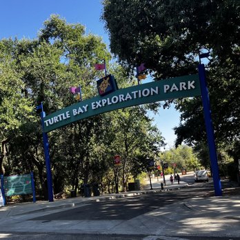Turtle Bay Exploration Park - Arch sign from parking lot - The photo captures the entrance sign for Turtle Bay Exploration Park, featuring a turtle logo and the park's name. The sign is set against a backdrop of tall, leafy trees and a bright blue sky. In the background, there are a few visitors walking and some cars parked or driving nearby. The scene is well-lit with natural sunlight, and the area appears to be part of a parking lot or driveway leading into the park. - Museums Near Me - Redding, California