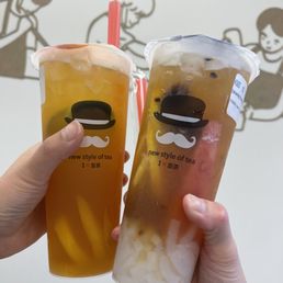 Mango Passion - Two people hold cups of tea with lemon slices and fruit pieces, with a cartoon mural of children in the background.