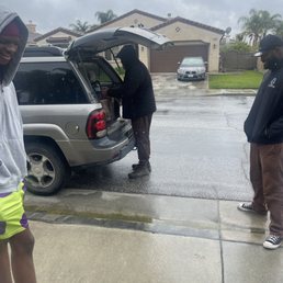 Checking key - Three men are positioned around a silver SUV parked on a wet street in a residential area. One man is loading or unloading items from the open trunk, while the other two stand nearby, observing. The scene is set on a rainy day, with overcast skies and wet pavement. The background features houses with garages, a parked car, and some palm trees, indicating a suburban neighborhood.