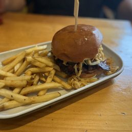 Alabama Slamma - A burger with a shiny bun, shredded cheese, and bacon is presented on a white oval plate. The burger appears to have additional toppings and is accompanied by a portion of French fries. The setting is a casual restaurant with a wooden table, and blurred background elements suggest a relaxed dining environment.