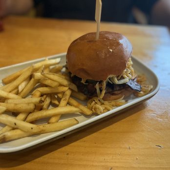 Thirsty Beaver - Alabama Slamma - A burger with a shiny bun, shredded cheese, and bacon is presented on a white oval plate. The burger appears to have additional toppings and is accompanied by a portion of French fries. The setting is a casual restaurant with a wooden table, and blurred background elements suggest a relaxed dining environment. - Burgers Near Me - Smithfield, Rhode Island