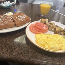 Breakfast Combo no meat - The image shows a breakfast setting with two plates of food and beverages on a dark brown countertop. One plate contains French toast topped with powdered sugar and a small serving of butter. The other plate has sliced tomatoes, scrambled eggs, and pieces of breakfast casserole or hash. There is a glass of orange juice and a glass of water nearby. In the background, part of a person’s arm and hand are visible, along with a small dish of creamers and a napkin dispenser.