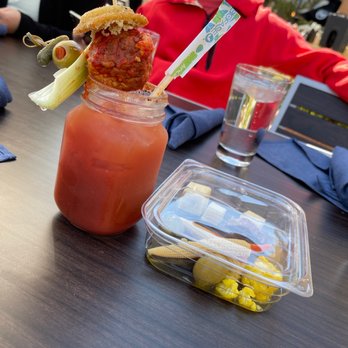 Hash Kitchen - Blood Mary! - A Bloody Mary cocktail is presented in a glass jar, garnished with a pickle, celery stalk, and a fried chicken drumstick skewered on a stick. The drink has a reddish hue, typical of tomato juice-based cocktails. The setting appears to be a restaurant or outdoor dining area, with other drinks and food containers visible on the table. A person in a red jacket is seated nearby, and the table has blue napkins and a plastic container with food. - Breakfast & Brunch Near Me - Chandler, Arizona
