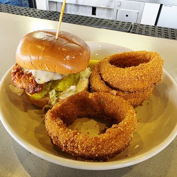 Showroom - Nashville Hot Chicken Sandwich with a side of onion rings - A white round plate contains a chicken sandwich with a bun, fried chicken fillet, pickles, lettuce, and melted cheese. The sandwich has a toothpick inserted into the top bun. Next to the sandwich are three large, golden-brown breaded onion rings. The plate is placed on a light-colored table with a textured black and white surface in the background. - New American Near Me - Frederick, Maryland