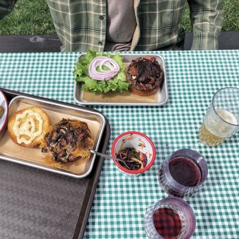 James Ranch Market - A table with a green and white checkered tablecloth displays multiple food items including a plate of potato wedges, a dish with a baked item topped with mayonnaise, a small bowl of salad, and a tray with a burger topped with onions and lettuce. There are three glasses of beverages, including red wine, beer, and a light-colored drink. A person wearing a green plaid shirt is seated at the table, with only their torso visible. - Farmers Market Near Me - Durango, Colorado